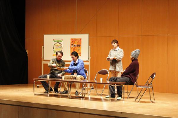 舞台の上の長机に座っている男性3名とお盆を持ってお茶を配ろうとしている割烹着姿の女性の写真