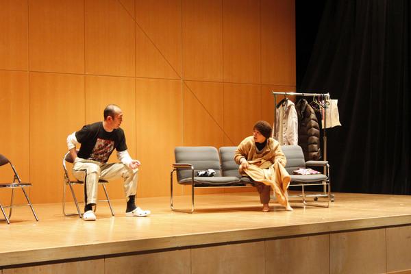 舞台の上でパイプ椅子に腰かけた男性と帽子を被った男性がソファーに腰かけて話をしている写真