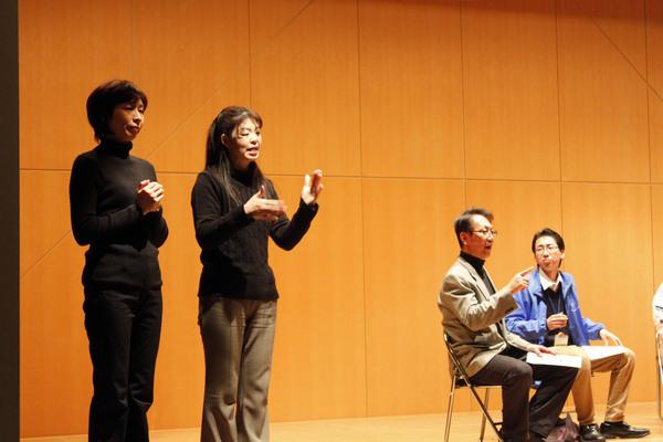 舞台の上で男性2名が椅子に座って話している様子を手話通訳している女性とその横に女性1名が立っている写真