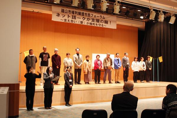 舞台の上に劇の出演者が並んでおり、舞台下でマイクで女性が話しているのを手話通訳している女性と横で立っている女性の写真