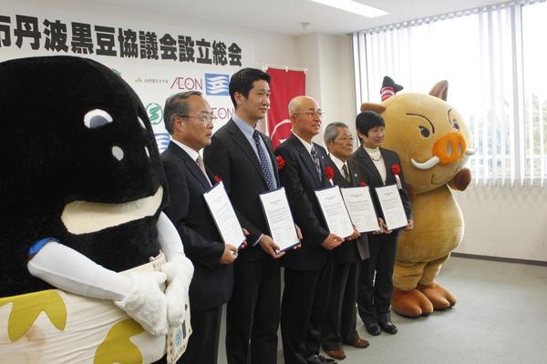 イメージキャラクターと市長と関係者で書類を持ち前を見ている姿の横から見た写真