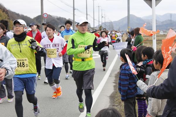 道路で一生懸命走っている選手に子供達が旗を振りながら応援している写真