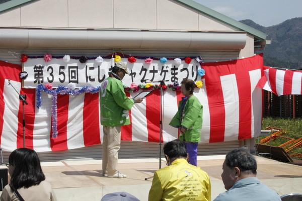 「第35回にしきシャクナゲまつり」と書かれている横断幕の前で緑の法被を着た女性が表彰されており、男性が賞状を読んでいる様子の写真