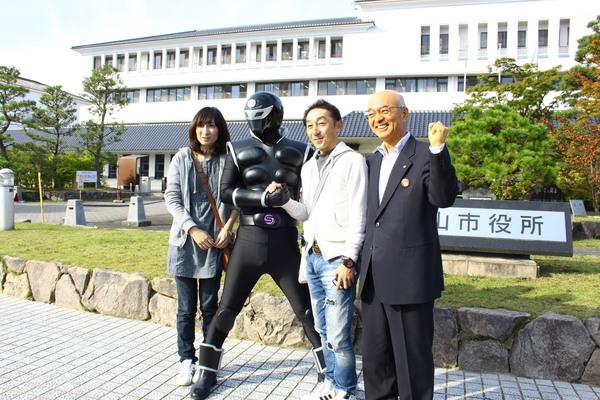 神戸から訪れた男女の観光客とササグローが握手をして市役所の前で市長も交え記念撮影している写真