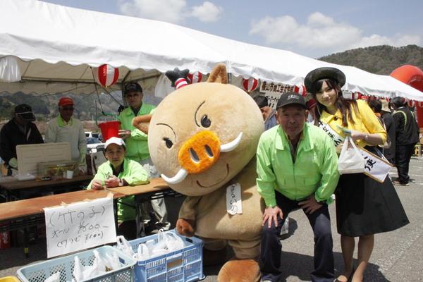 お祭り会場の出店の前で観光大使の女性と黄緑色のジャンパーを着た男性と出店者と「まるいの」が一緒に写っている写真