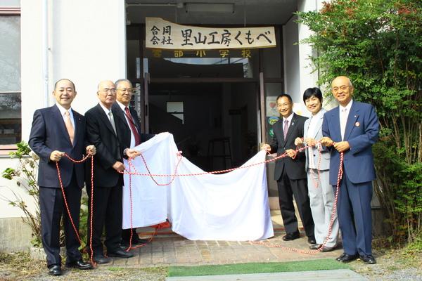 市長及び関係者が紅白のロープと白い布をもっており、市長が書いた「合同会社 里山工房くもべ」の除幕式の様子の写真