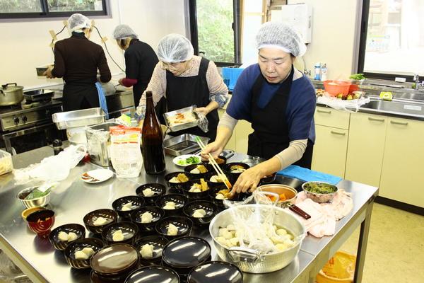 調理室でヘアキャップをかぶり、黒いエプロンを付けた女性が、台に並べられたお椀に里いもやカボチャを綺麗に盛り付けしており、後ろにも料理をしてる女性が写っている写真