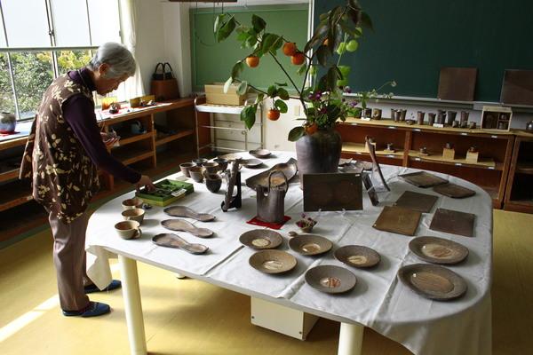 教室の一室に、陶芸品が沢山置かれており、テーブルの中央に柿の実がなった枝が壺に生けてあり、周りにはコップや色々な形のお皿が置かれており、年配の女性が焼物を見ている写真