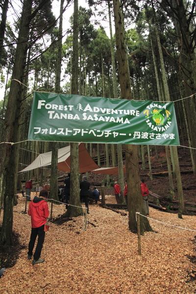 杉林の中に、「フォレストアドベンチャー・丹波ささやま」と書かれた看板が木に結び付けられ、奥の方では木々に結び付けらた布で屋根が作られており、その下に椅子に関係者が座っている様子の写真