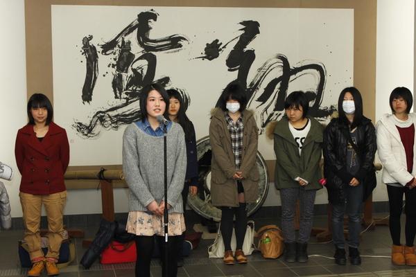 後ろの壁に大きな文字で描かれている作品の前で女子高生が代表でお話ししている写真