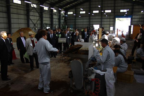 作業着姿の男性がシャベルを持って、灰色の機械の中に土を入れようとしている写真