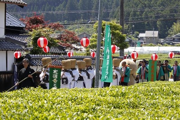 沿道に提灯が飾られ、白い衣装と天蓋の帽子を被り、木の笛を吹いた方々の後ろを法被を着た人々が並んで歩いている写真