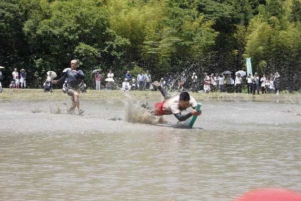 田んぼの中で、緑色の棒を取った男性と走っている男性がおり、その様子を見ている人々の写真