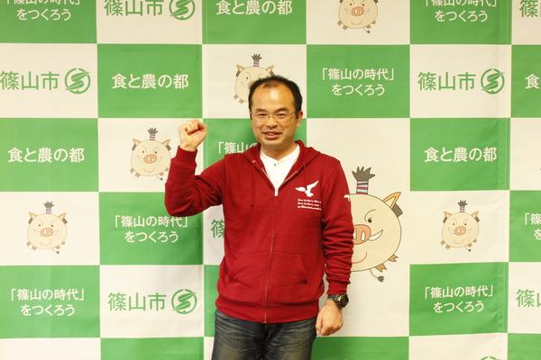 赤色のパーカーと黒のジーンズ姿の酒井 正幸主査が右手でガッツポーズをしている写真