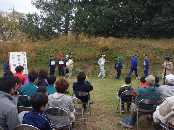 頭に緑や、赤、茶色の生物のお面をかぶり、観客の前で劇をする生徒の写真