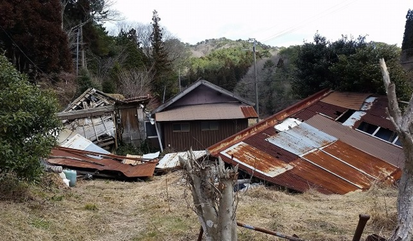老朽化で傾いた空き家と錆のついた廃材が散乱している写真