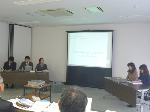 報告会でスクリーンを使って男性の方が説明しているのを他の職員の方々が資料をみて聞いている写真