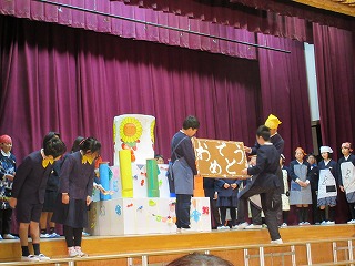 紙で作られた大きなバースデーケーキの前で、おめでとうと書かれたボードを3人の生徒が持ち、それを見る舞台上の生徒たちの写真