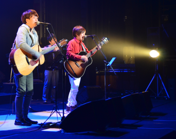 ギターを弾きながら歌を歌っている兄弟デュオ「ちめいど」の2人に照明の光が当たっている写真