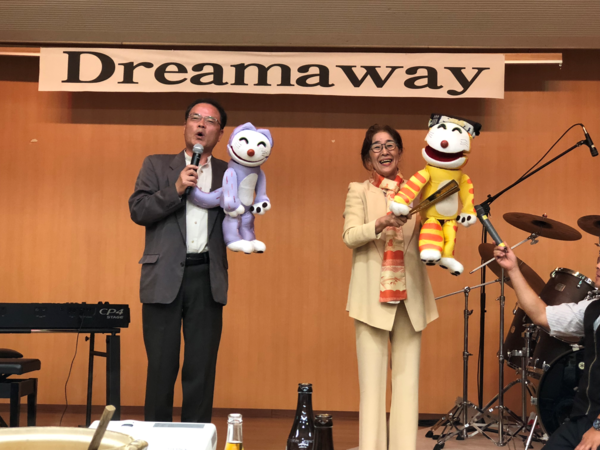 舞台の上で、人形を持ち発言をしている男性と笑顔の女性の写真