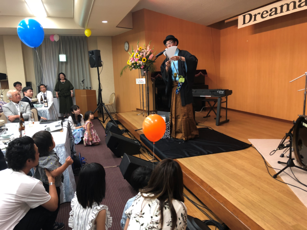 舞台上、子供たちが集まる前で紙芸をしている男性の写真