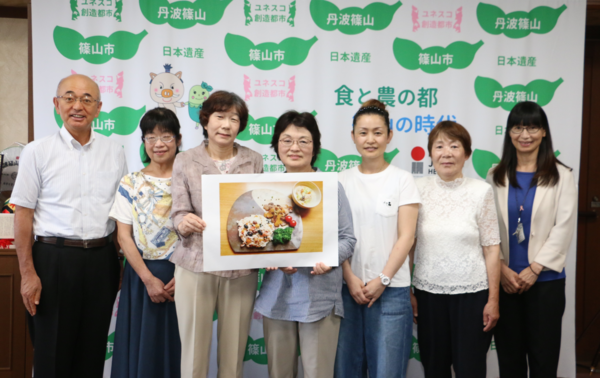 篠山市地域活動栄養士会の皆さんが料理の写真を持ち、市長と集合写真
