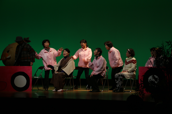 舞台の上に4名の女性たちが座っていて、その後ろにピンクのジャンバー姿の女性たちが3名立っている写真