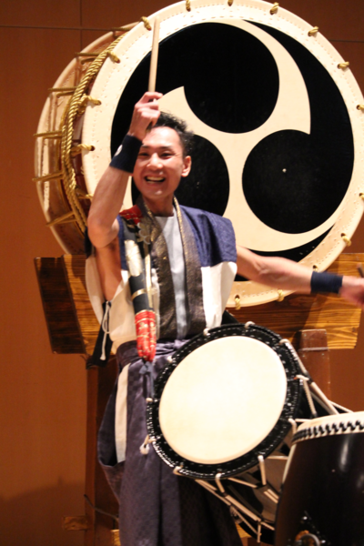 男性が笑顔で肩に下げた太鼓をたたいている写真