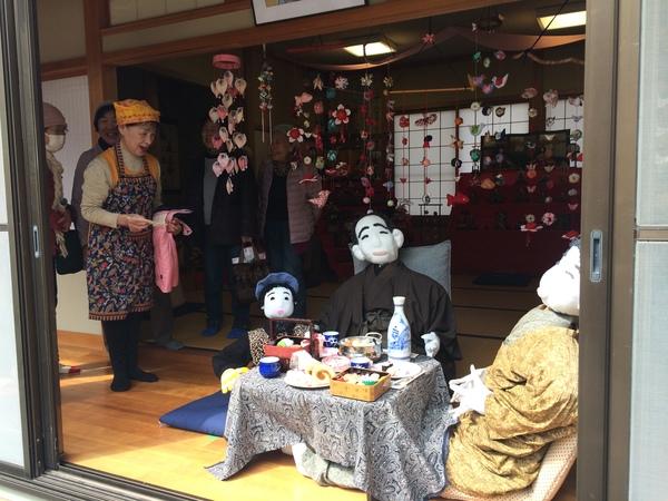 民家の縁側で、机の上に並べられたご馳走を囲んで座る、かかしの家族の写真