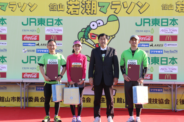 梛野 泰子さん、小西 隆紀さん、俣野 秀明さんが賞状と記念品を持って主催者の男性と一緒に記念撮影している写真