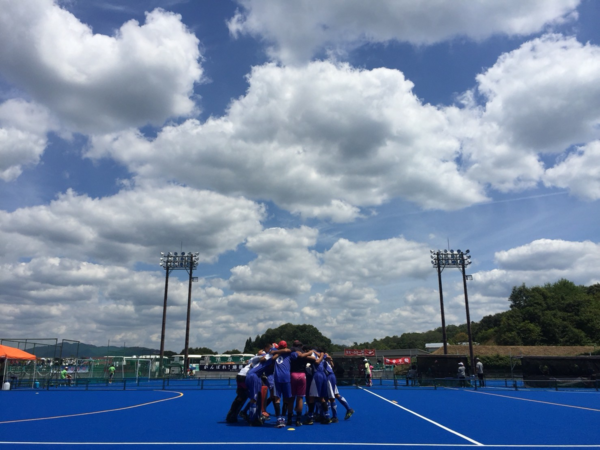 雲の多い青空の下でユニフォームを着た選手たちが中央で円陣を組んでいる写真