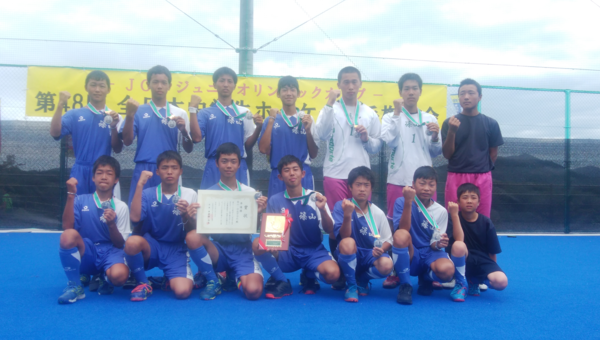 12名の選手が首にメダルをかけ、中央の選手が盾と賞状を持ってガッツポーズをしている写真