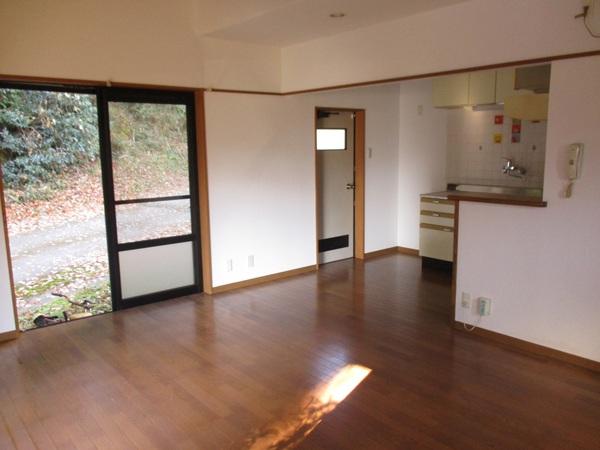 フローリングの床で窓が開けられていて外の道が見えて奥に台所がある部屋の写真