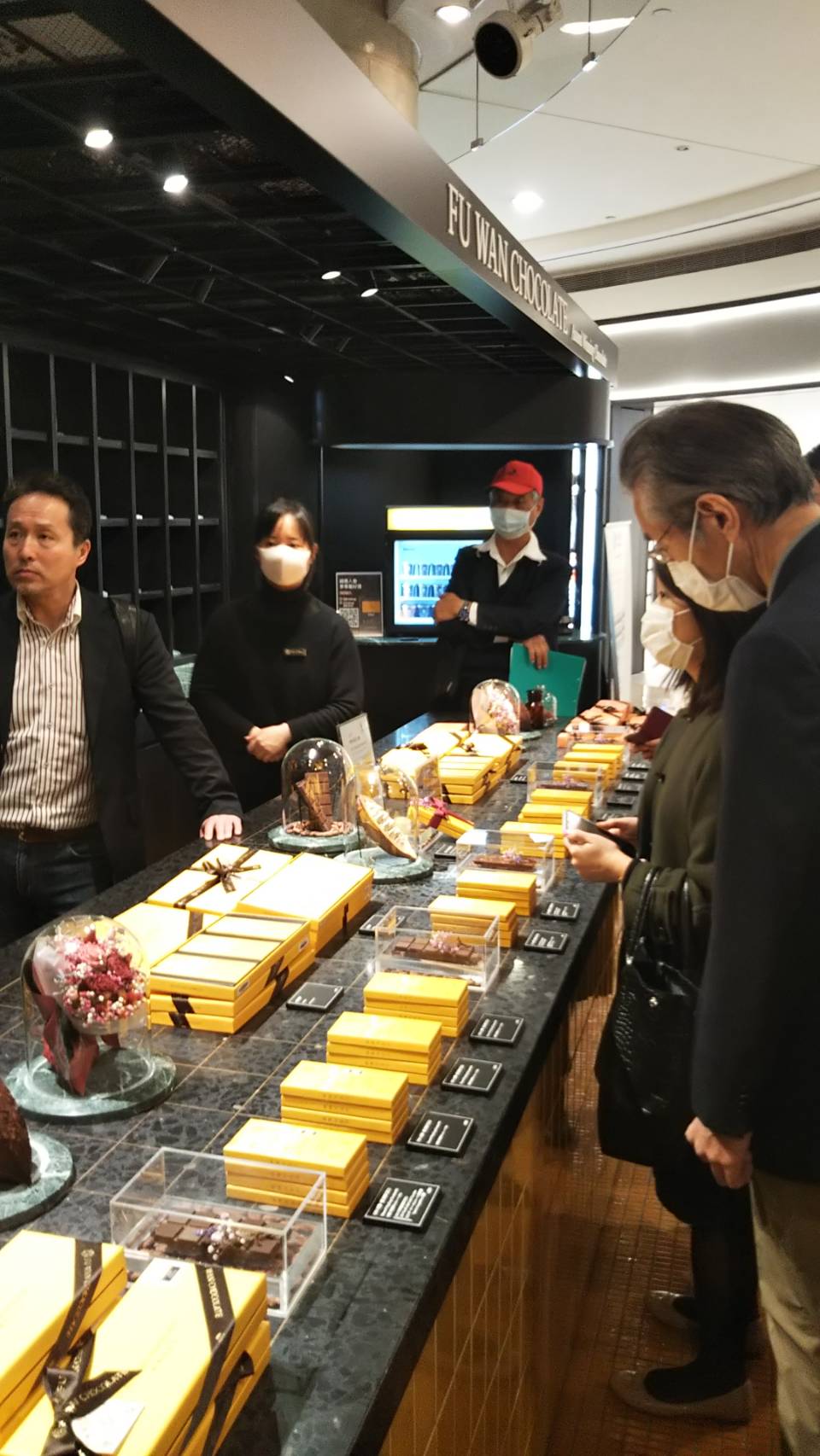 フ―ワンチョコレートを販売しているお店で、机の上に等間隔で黄色の箱に入ったチョコレートが並んでおり、その周りを囲んでチョコレートを見ている人々の写真