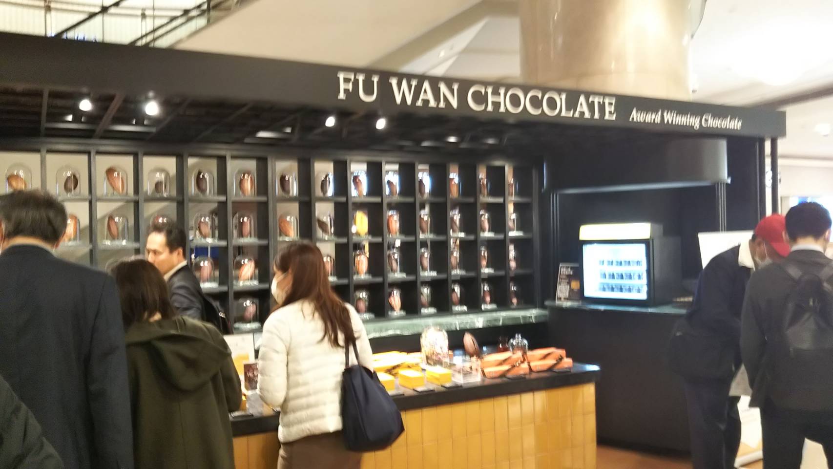 フ―ワンチョコレートを販売しているお店で、チョコレートを見る人々と、区切られた棚に瓶に入ったカカオのようなものが飾られているのが分かるお店の全体の写真