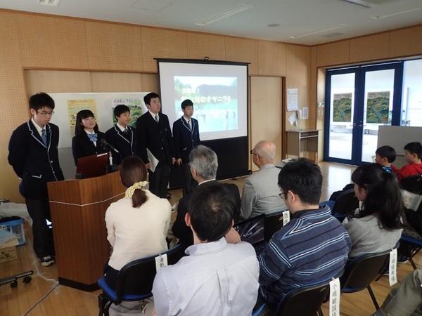 制服を着た高校生5名がプロジェクターで映像を写しながら発表をしており、出席者が椅子に座って話を聞いている様子の写真