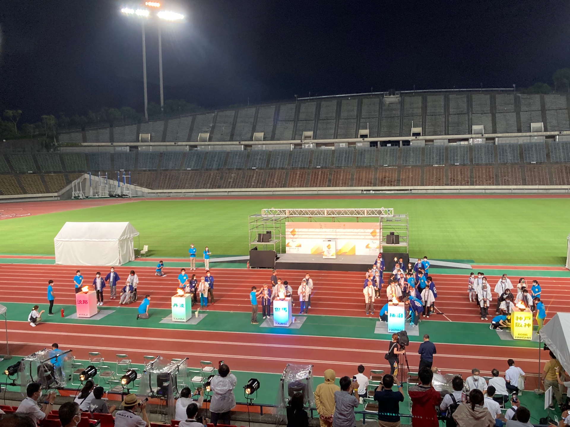 スタジアムに、聖火を集めるための台が5つつ並べられており、まわりに関係者が立っている