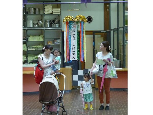 子どもを連れた女性が二人立っていて、その間に「祝ちるみゅー入館一万人」と書かれたくす玉が割られている写真