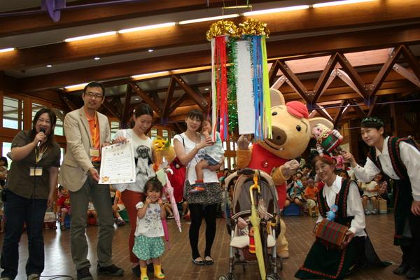 くす玉の周りにマイクを持った女性や賞状を持った男性、子どもを抱っこしている女性、ぶたの着ぐるみなどが並んでいる写真