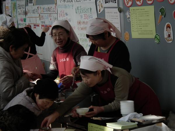 三角巾にエプロン姿の女性たちが客に食べ物を振舞っている写真