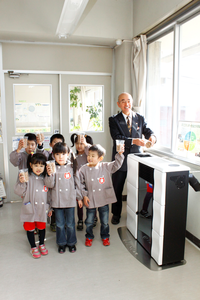 灰色のスモッグを着た園児たちが、透明なコップに入った木質バイオマスを持っており、市長が四角い機械にコップに入った木質バイオマス入れようとしている様子の写真