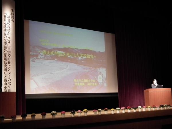 篠山の特色を生かした学校給食について、栄養教諭 岡田氏がスクリーンをバックに舞台で話している様子