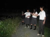 暗くなった川岸に制服を着た高校生が集まっており、ホタルを見つけて指差ししている様子の写真