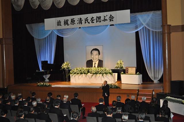 舞台の上に笑顔で写っている梶原 清さんの遺影と白色の花が置かれ、ステージ下の椅子には沢山の参加者が座っており、1人の男性がマイクを持ちながら立ち、話をしている写真