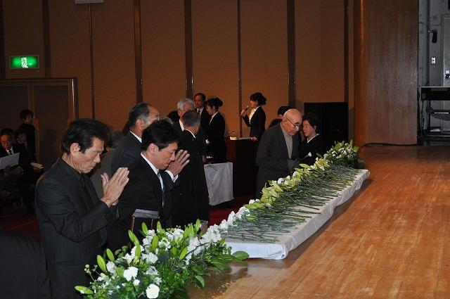 ステージ上に献花台が準備され、参加者が白色のカーネーションやユリの花を置き、手を合わせている写真