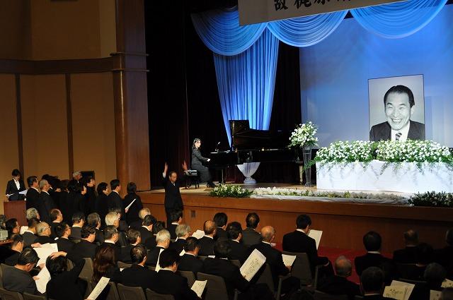 参加者がステージ上の梶原 清さんの遺影を前にピアノ伴奏と指揮者に合わせて歌を歌っている写真