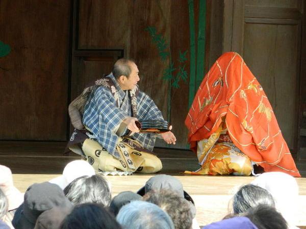 能舞台にて男性が赤い羽織を被った着物姿の女性に話しかけている様子が演じられている写真