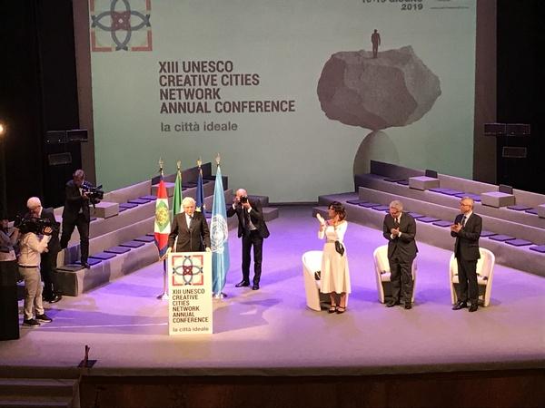 イタリア大統領が壇上で話をしているのを聞いている3名が立って拍手している様子をカメラマンが撮影している写真