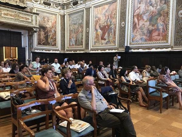 絵が飾られている会場で椅子に座って話を聞いている人々の写真