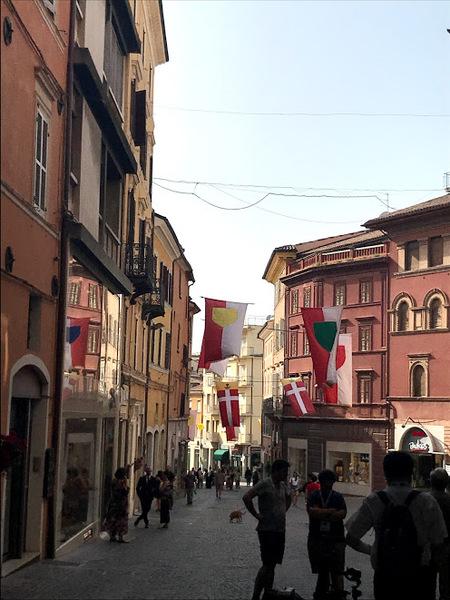 ピンクに近い色の外壁、真っ白の外壁の建物が並びところどころに飾られた旗がなびいているイタリアの街並みの写真